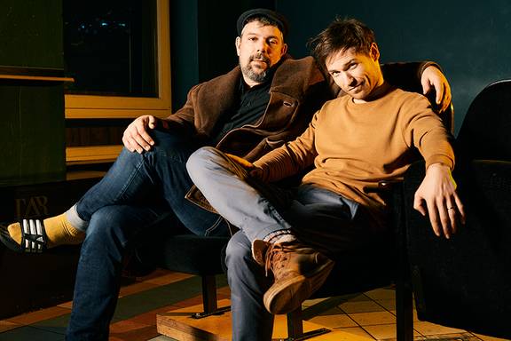 Felix Römer und Martin Bosch (Beschreibung: Zwei Männer schauen sitzend in die Kamera. Beide haben kurze dunkele Haare, einer mit Bart, der andere trägt ein ocker-farbenes Oberteil.)